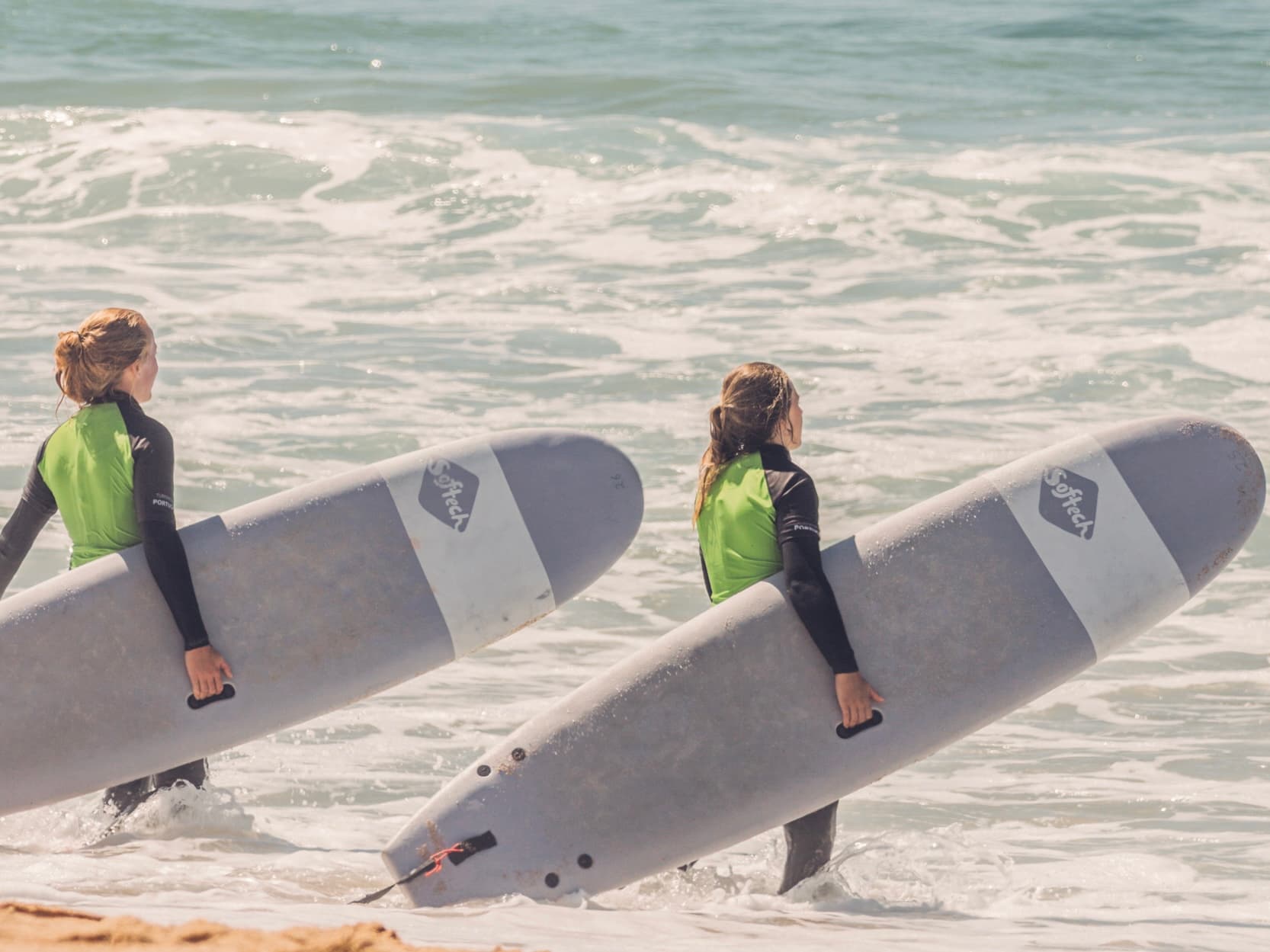 Zwei Surfschülerinnen mit ihren Surfboards unter dem Arm gehen ins Wasser